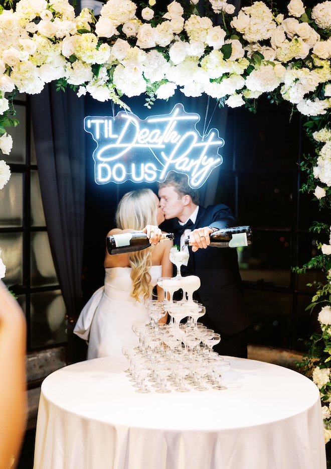 The bride and groom kissing while pouring champagne into a champagne tower with a sign that says "'Til Death Do Us Party". 
