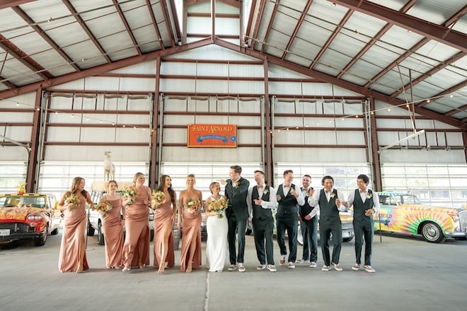 The couple and their bridal party walk beside antique decorated cars at Saint Arnold Brewery in Houston. 
