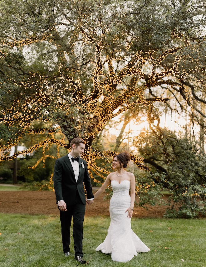 The bride and groom hold hands in front of an oak tree wrapped in twinkling lights at The Houstonian Hotel, Club and Spa. 