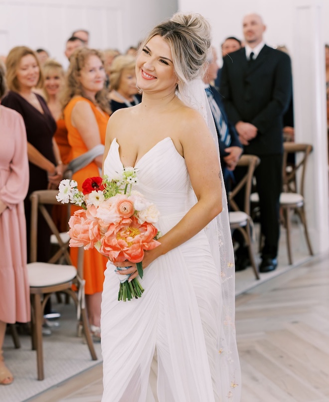 The bride holds an orange wedding bouquet as she walks down the aisle at The Pattison House.