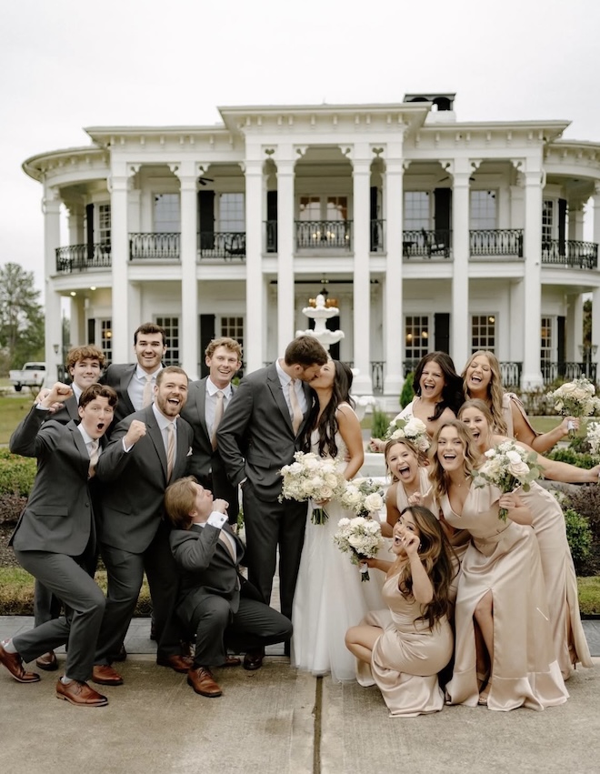The bride and groom share a kiss as the wedding party cheers around them outside of Sandlewood Manor.