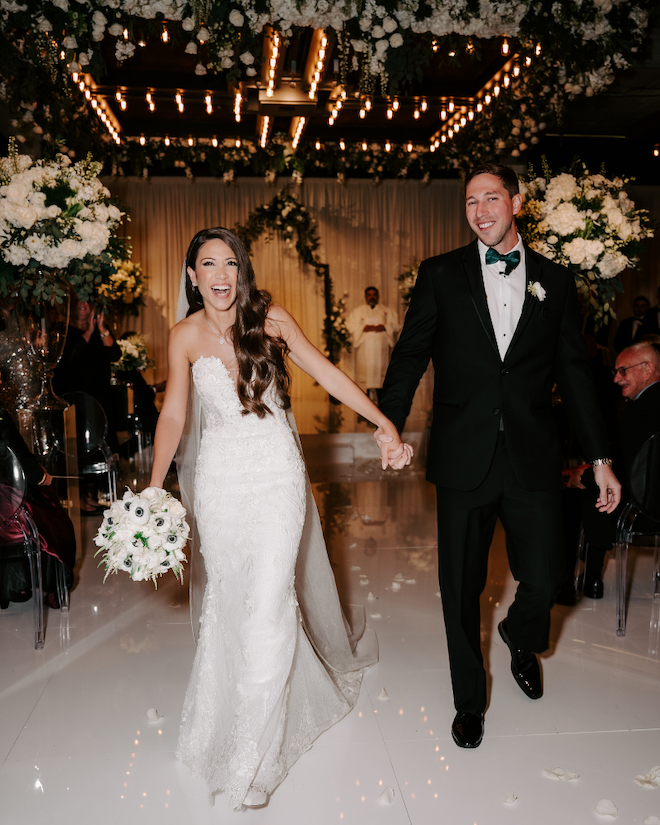 Bride and groom walking down aisle holding hands and smiling with green and white florals behind them.