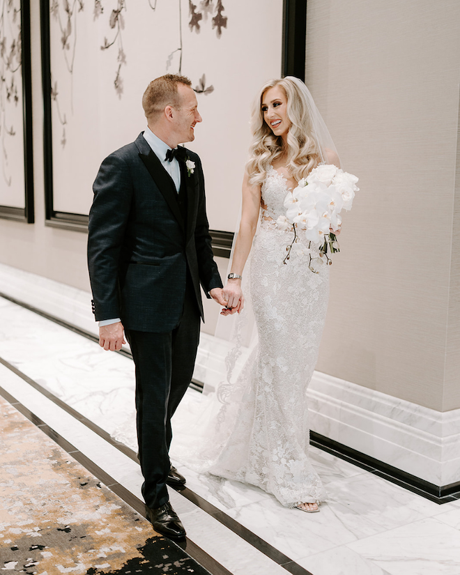 Bride and groom holding hands walking down hotel hallway smiling and staring at each other with a white bouquet in bride's hands/