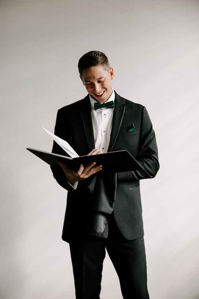 Groom in black suit with dark pine green bowtie reading note bride gave him.