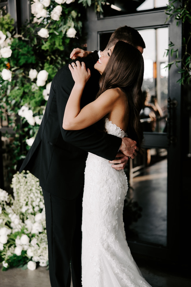 First look with the bride and groom, hugging and smiling surrounded by green and white florals.