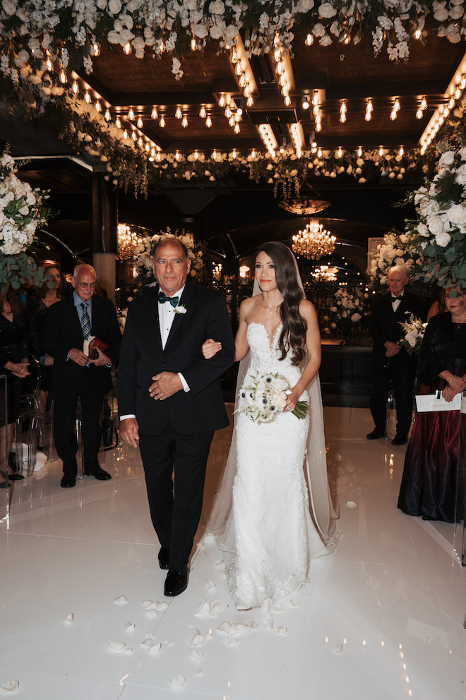 Bride and her father walking down the aisle with green and white florals around them.