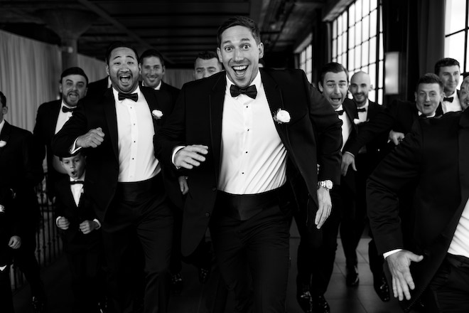 Black and white photo of the groom and his groomsmen running and smiling towards camera.