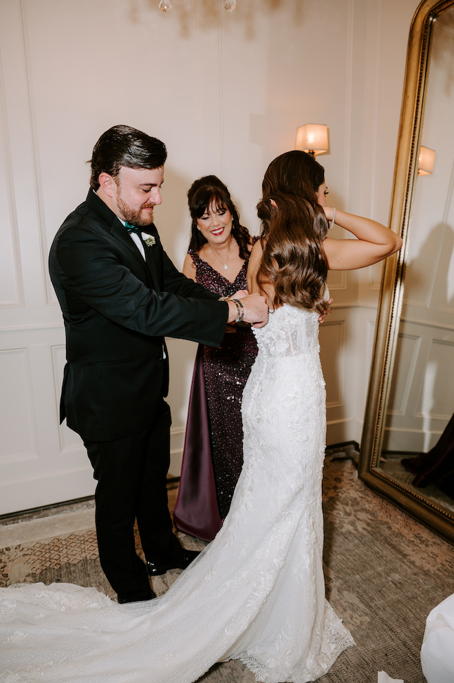 Bride's mother and her brother in law helping her put on gown in front of mirror.