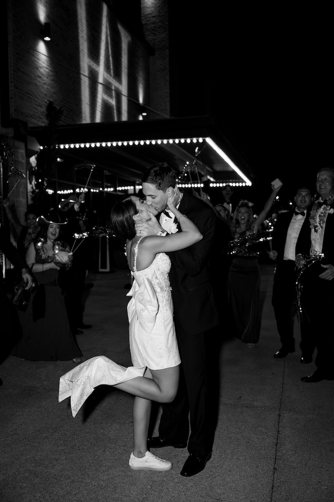 Black and white photo of the newlywed couple kissing during exit and guests cheering.