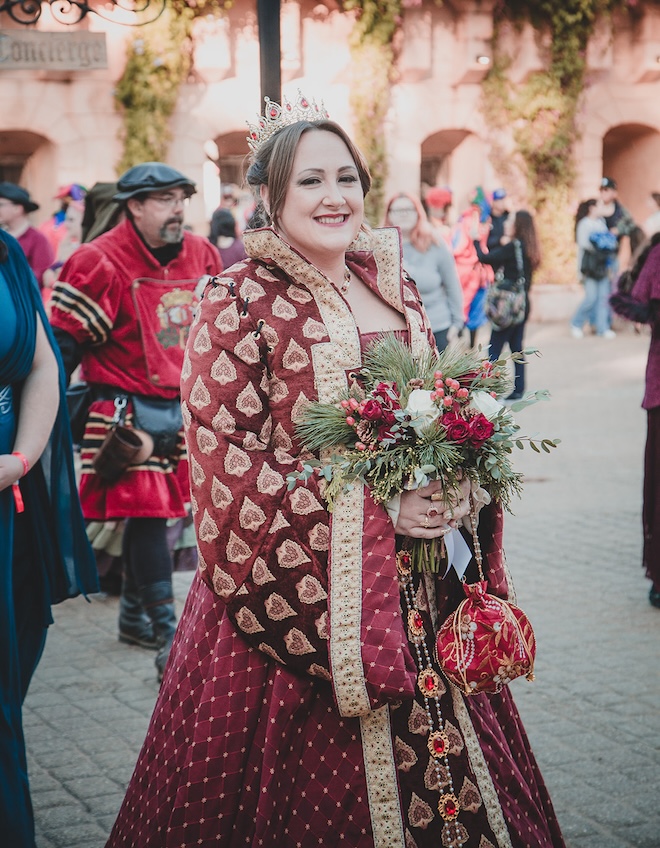 The bride wearing gold and burgundy holding a bouquet on the grounds of the Texas Renaissance Festival.