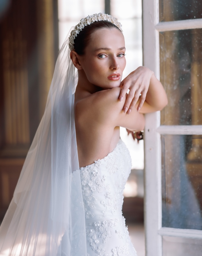 A bride leaning on a window wearing a floral headpiece and veil and a strapless wedding gown.