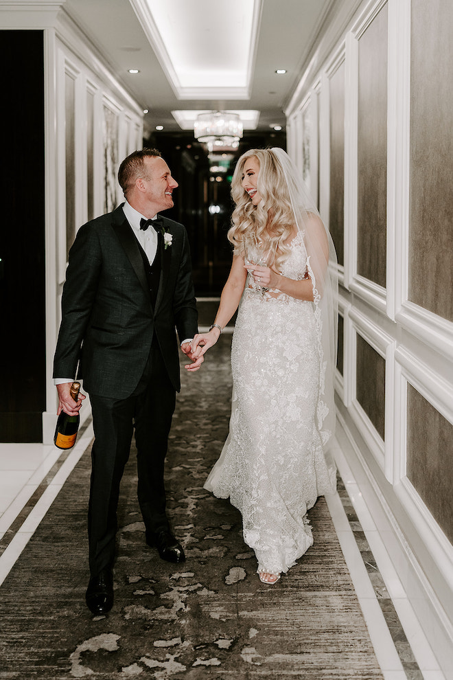 Bride and groom walking down hotel hallway smiling and laughing with each other with a champagne bottle and glasses.