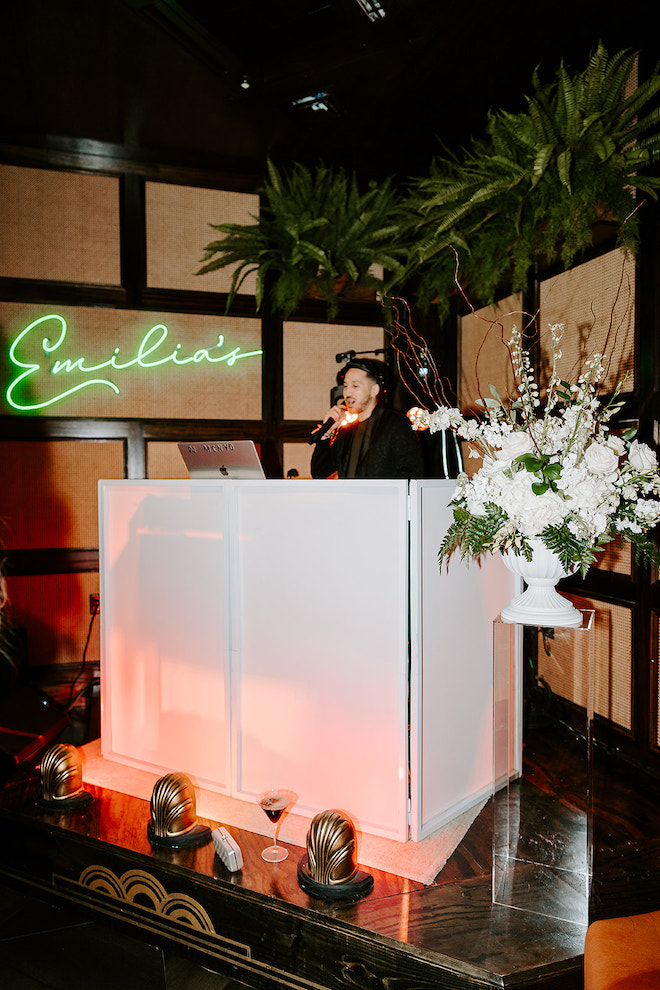 DJ at after-party at big white booth with neon green sign that read "Emilia's" with a white bouquet next to the booth.
