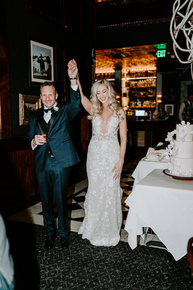 Newlywed couple walking into reception while holding their hands together in the air and smiling.