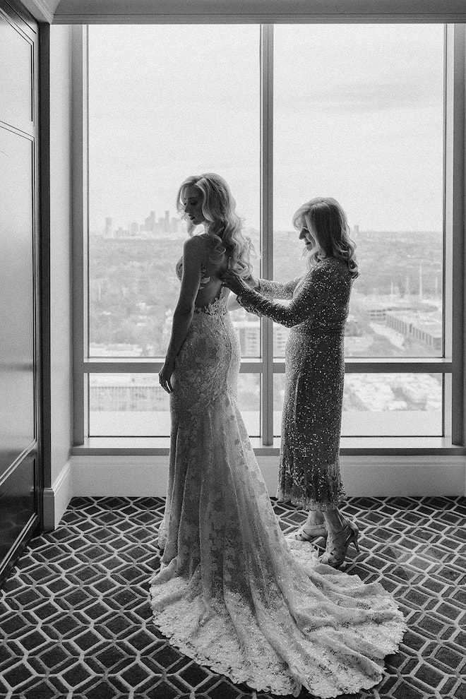 Black and white photo of the mother of the bride zipping her up into her dress in front of a window view of the city.