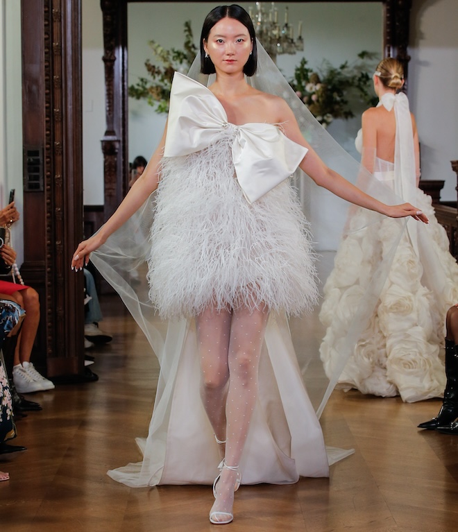 A model walking down the runway in a Monique Lhuillier ivory feather mini dress with silk Duchesse draped bow.