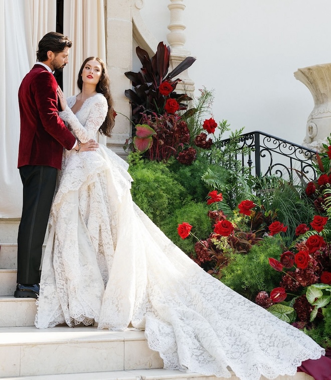 A bride and groom posing on a staircase with greenery and red florals behind them.