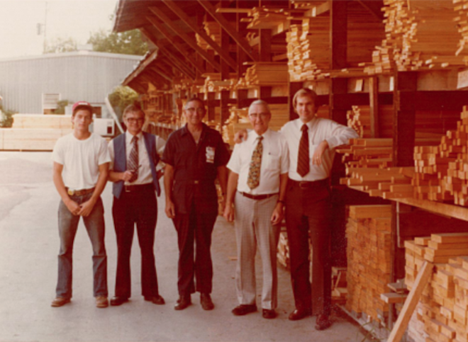 A historic photo of a group of men standing in fornt of plaques of wood. 