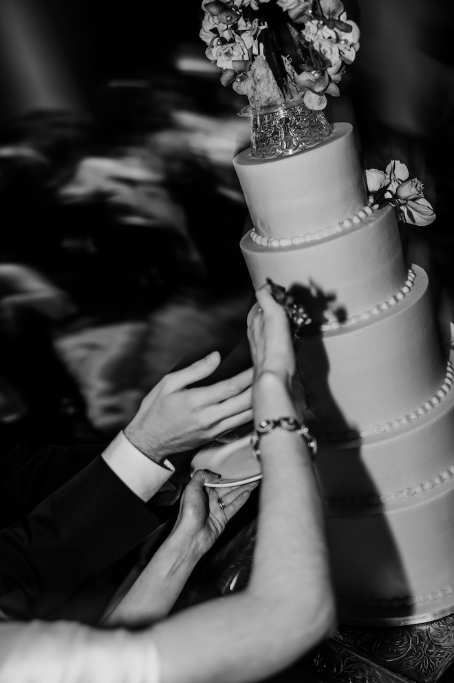 Black and white close up photo showing the couple cutting into a five tier white wedding cake. 