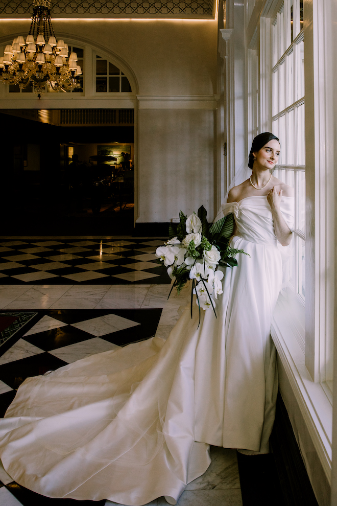Bride in wedding gown holding green and white bouquet looking out the window of the hallway in the venue.