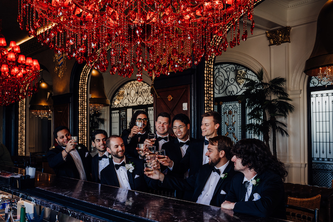 Groom and his groomsmen at the bar with a red chandelier above them cheersing with a drink.