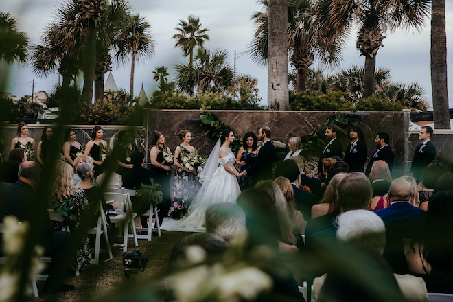 Farther view behind green and white plants showing bride and groom at the altar.