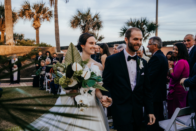 Bride and groom walking down the aisle holding hands and smiling while bride holds green and white bouquet.