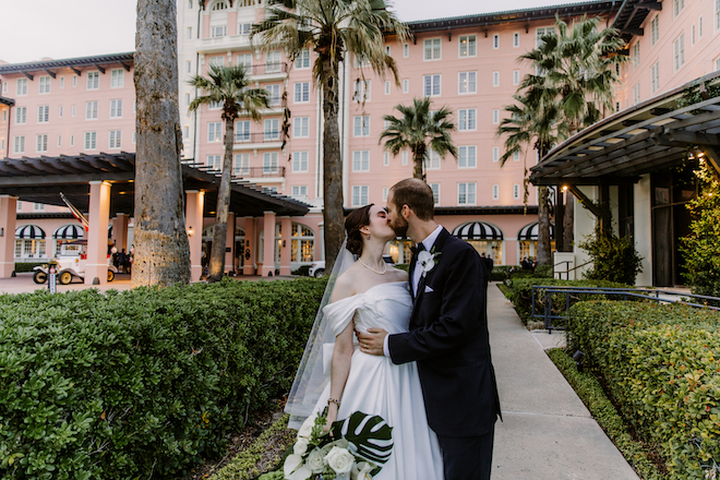 The couple kissing outside showing the venue behind them with tall palm trees.