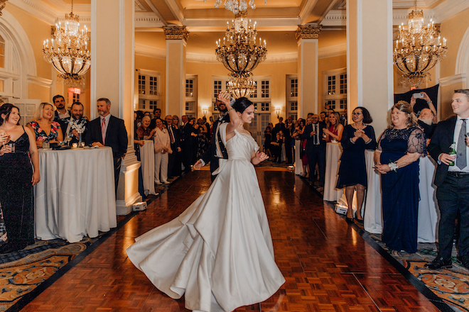 Bride and groom sharing their first dance while he spins her on the dance floor with chandeliers and guests around them.