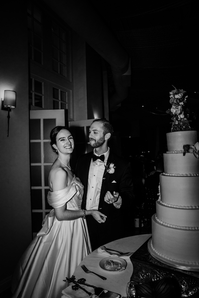 Black and white photo of the groom looking at the bride and both smiling before cutting into five tier white wedding cake.