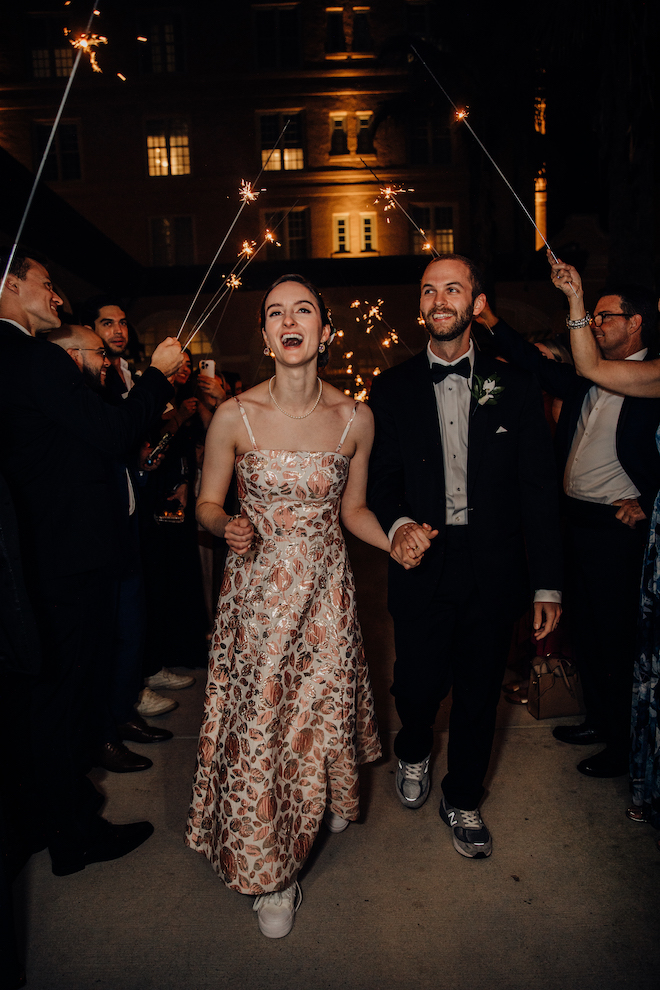 The couple exiting the venue with guests holding sparklers around them while they smile and hold hands.