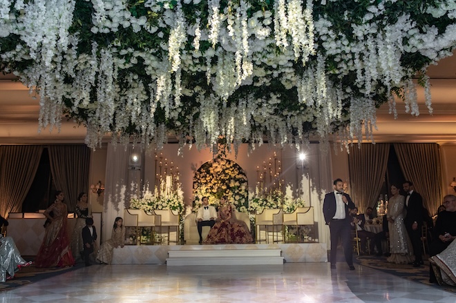A bride and groom sitting while a man gives a toast on the dancefloor under a canopy of white florals and greenery. 