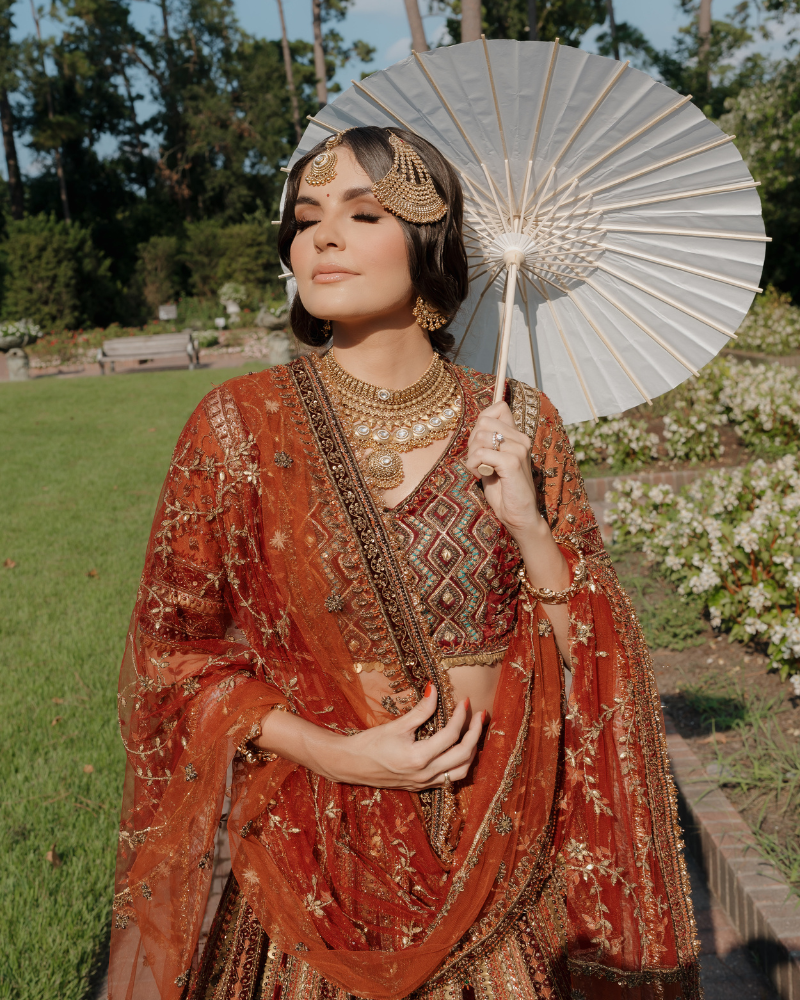 Model in red and orange toned traditional indian wedding dress in a green garden with hairpieces and holding an oil fan umbrella. 