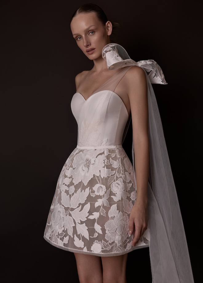 A bride wearing a Mira Zwillinger short couture gown featuring a strapless sweetheart corset crafted from duchess satin. The structured bodice flows into a floral embroidered skirt. The look is elevated by a detachable tulle topper with a dramatic bow.