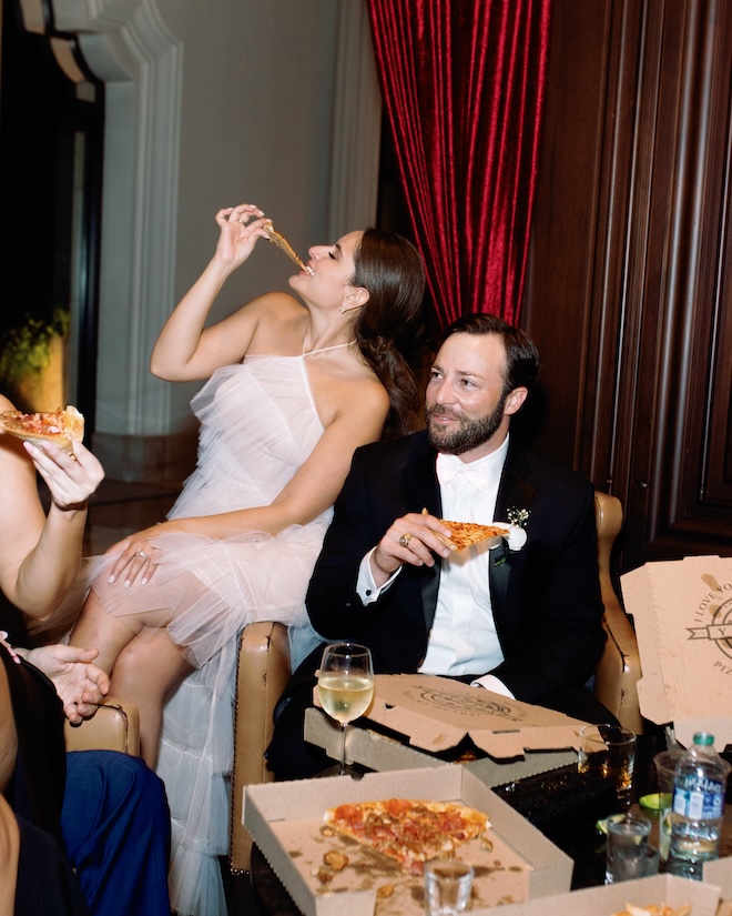 The bride and groom bite into pizza at their wedding reception.