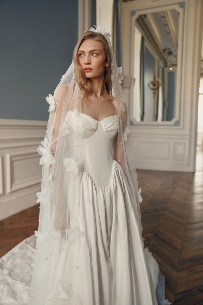 Bride wearing a Lihi Hod gown with a corset top and a basque waist paired with a veil with floral appliques. 