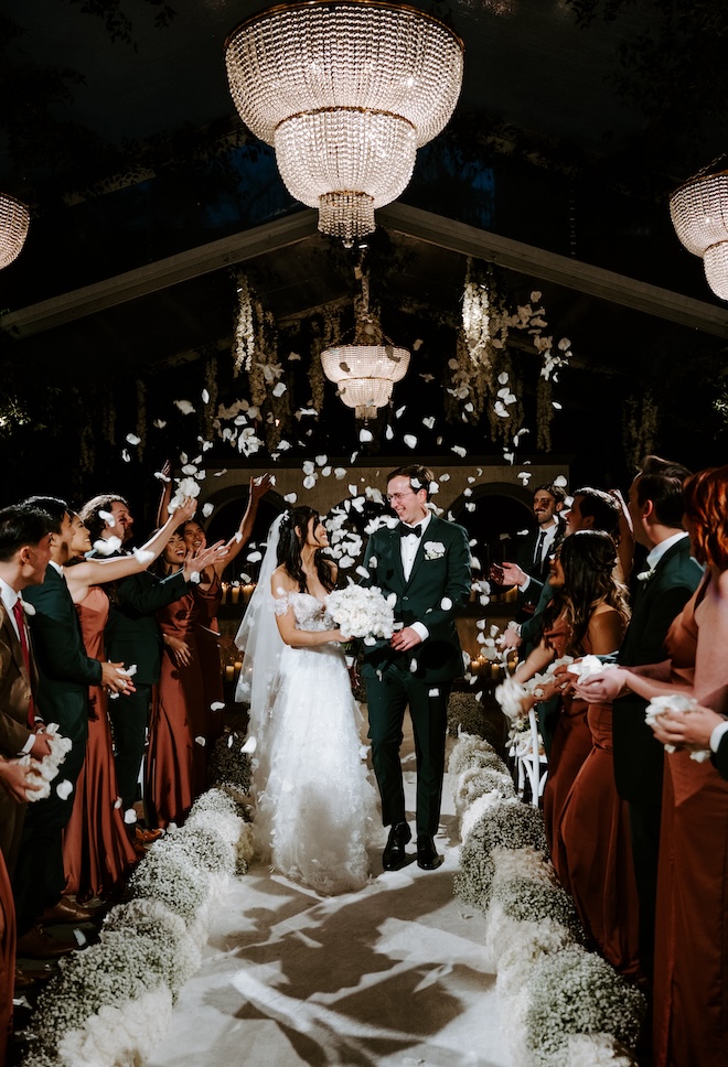 A bride and groom smiling at each other while walking down the aisle with guests throwing white petals around them.
