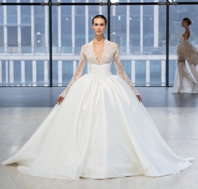 Model walking down the runway in a Nicole + Felicia Couture ballgown with a basque waist and a long sleeve lace top. 