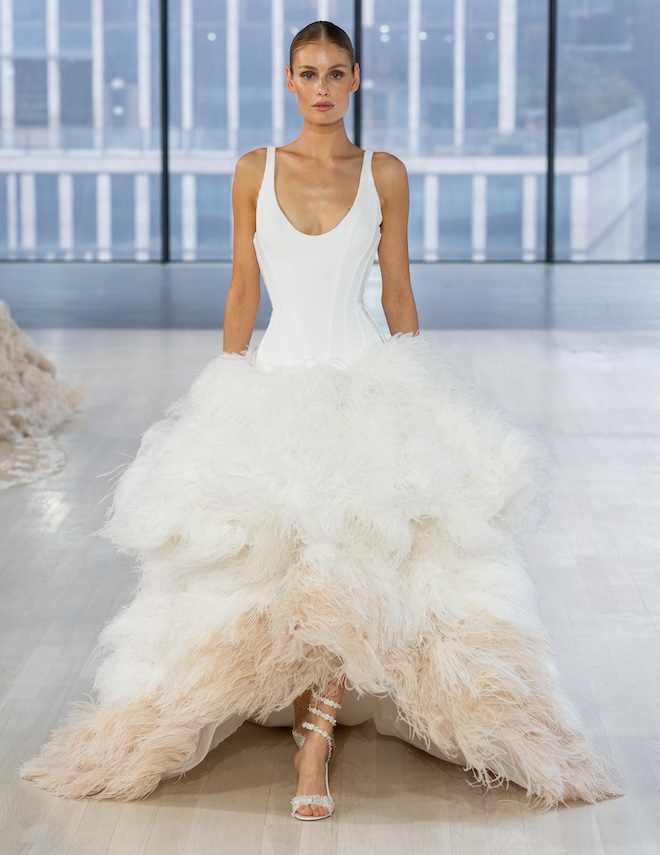 Model walking down the runway in a Nicole + Felicia Couture in a gown with a feathered skirt. 