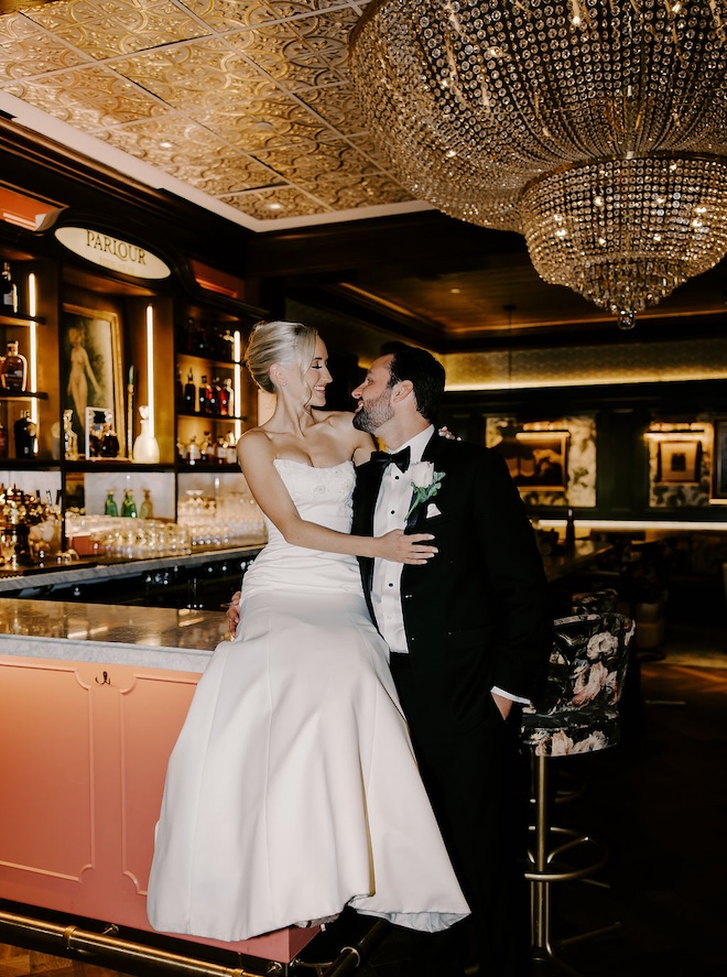 A bride and groom smiling in front of a bar at the C. Baldwin that is decorated with deep colors. 