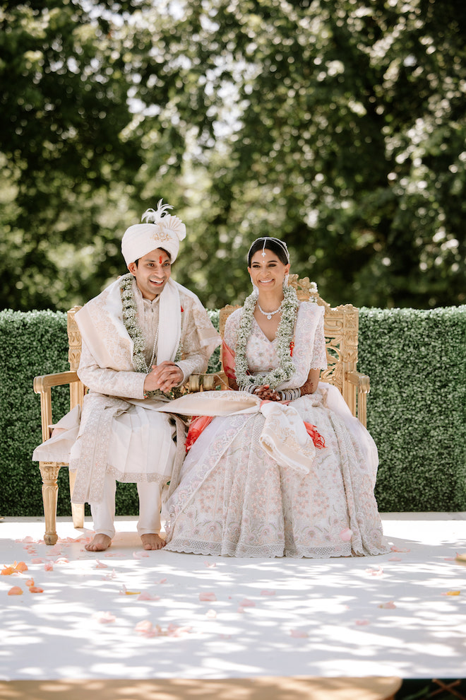 Bride and groom sitting in chairs doing traditional Hindu ceremony with large lush necklaces around their necks while they smile.