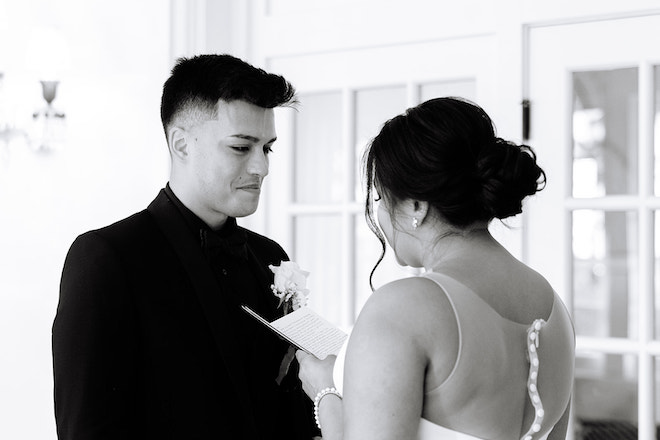 Black and white photo of the bride reads her vows and the groom is smiling looking at her.