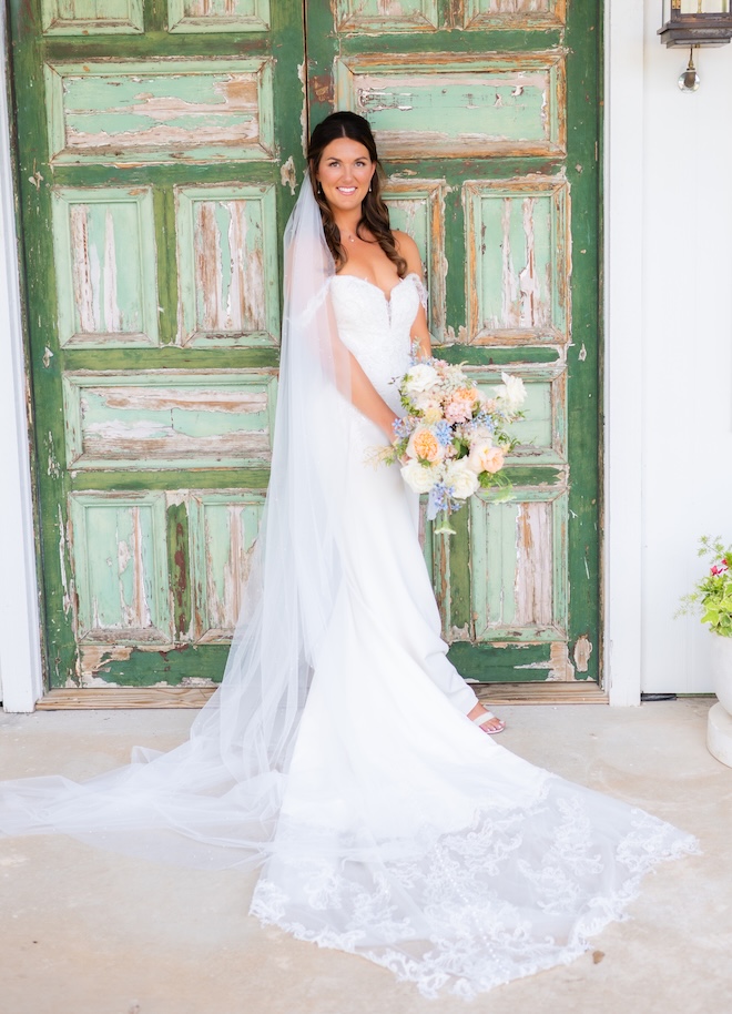 The bride stands in front of a green old barn door holding her wedding bouquet. 
