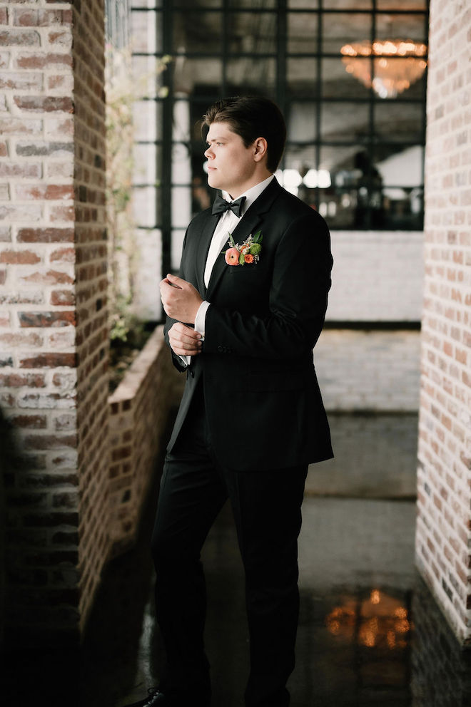Groom fixing cuff and looking out the window, in brick hallway of venue.