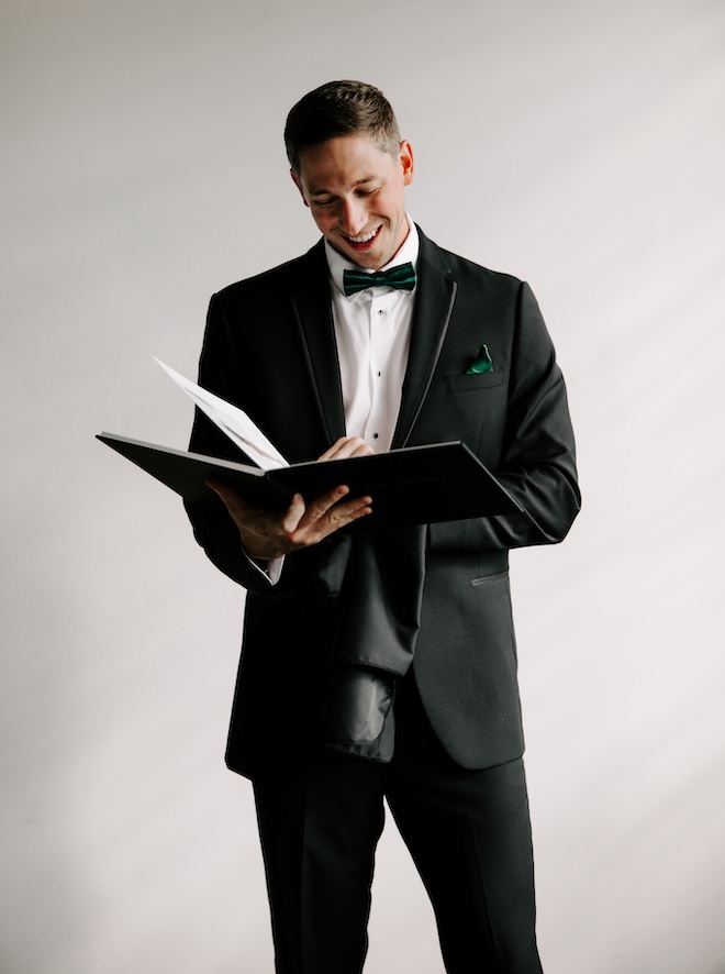 The groom reads a book before his wedding ceremony wearing his tuxedo with an emerald green bow tie and pocket square.