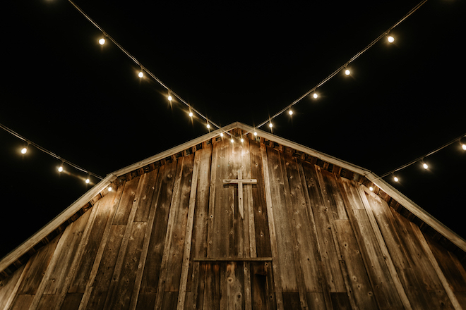 Outside of wooden barn chapel with string lights and wooden cross.