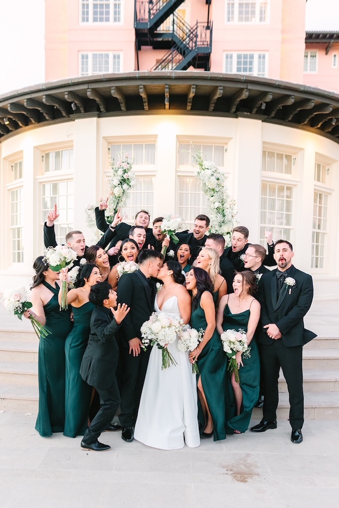 Bridesmaids and groomsmen surrounding the couple while they kiss and the wedding party cheers.