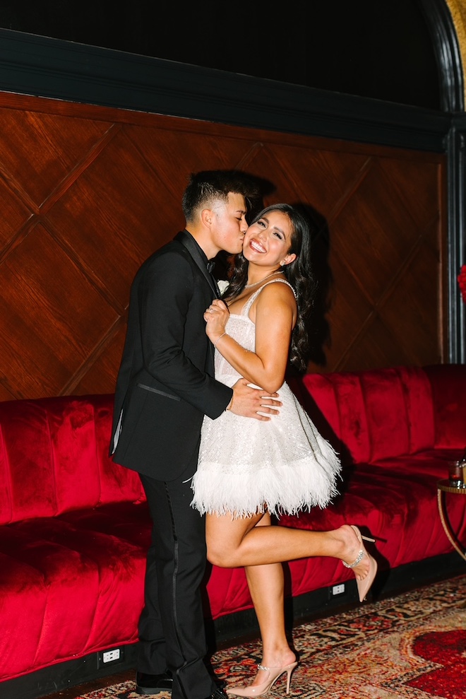 Groom kissing the bride on the cheek in front of red velvet couch.