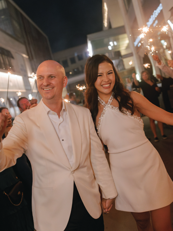 A bride and groom holding hands and smiling while guests wave sparklers behind them. 