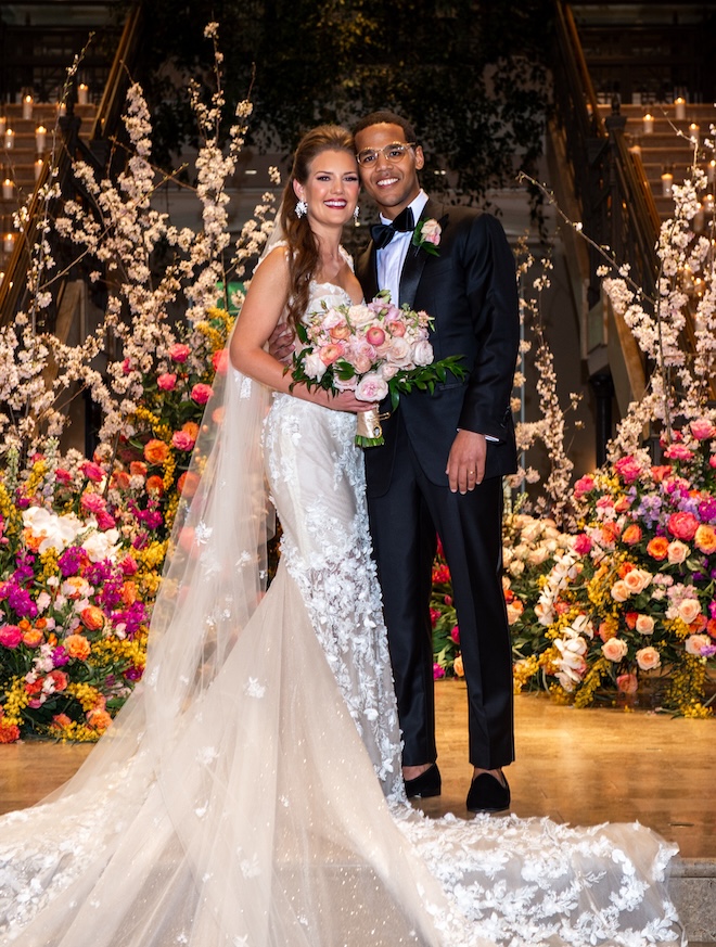 The bride and groom posing for a photo in front of a large and colorful floral installation by Plants N' Petals. 